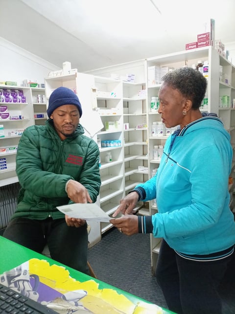 Man in green jacket and woman in blue jacket discussing paperwork inside a pharmacy with shelves of medications in background